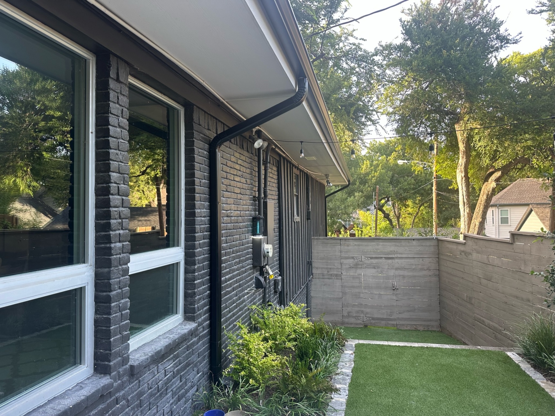 Gutter installation on a modern brick home in Lakewood, TX, showing sleek black downspouts, a landscaped backyard, and privacy fencing with turf lawn